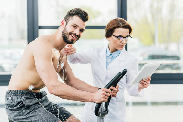 doctor with digital tablet near smiling shirtless sportsman during endurance test - Photo, Image