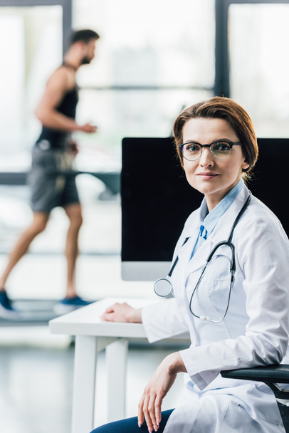 beautiful doctor sitting at computer desk in gym and looking at camera  - Foto, Imagem
