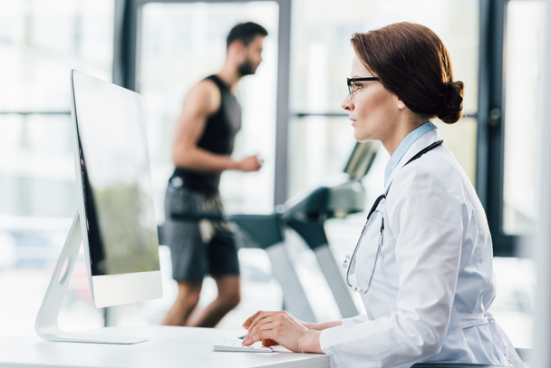 doctor in glasses using computer while sportsman running on treadmill during endurance test in gym - Fotoğraf, Görsel