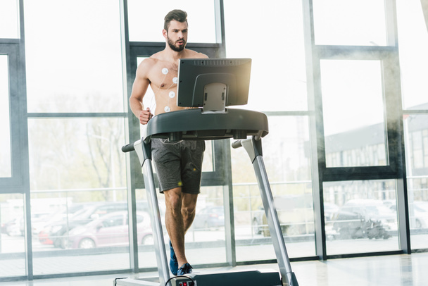 przystojny sportowiec z elektrodami biegowymi na bieżni podczas testu wytrzymałościowego w siłowni z oświetleniem dziennym - Zdjęcie, obraz