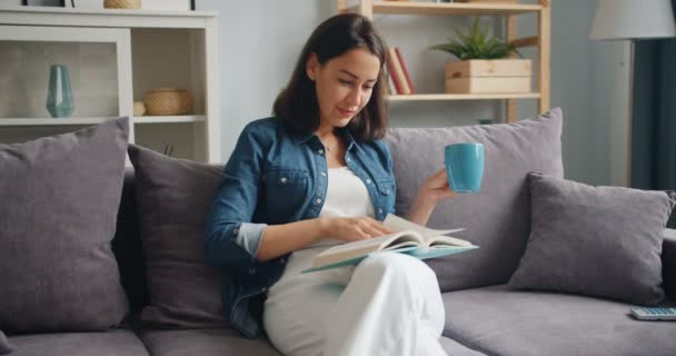 Cute student reading book and drinking coffee relaxing at home on sofa - Filmmaterial, Video
