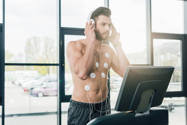 muscular sportsman in headphones running on treadmill during endurance test in gym - Photo, Image