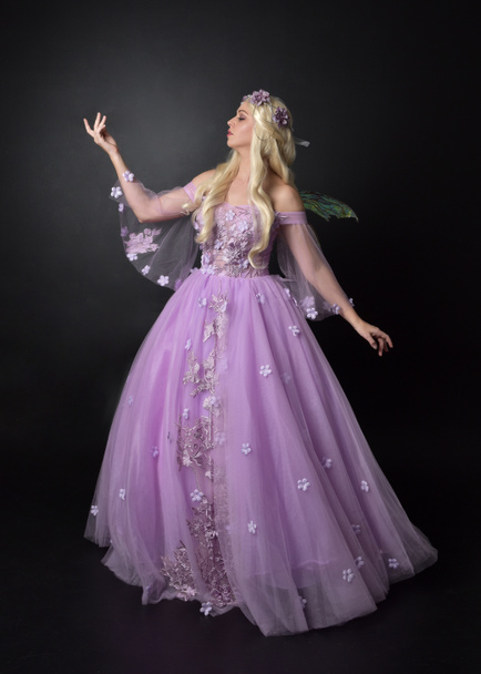 portrét blonďaté dívky v kostýmech s fantazií, dlouhé purpurové kuličkové šaty s víly, stojící póza na tmavém studiovém pozadí. - Fotografie, Obrázek