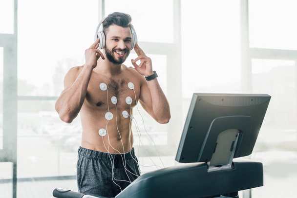 uśmiechnięty muskularny sportowiec w słuchawkach działających na bieżni podczas testu wytrzymałościowego w siłowni - Zdjęcie, obraz