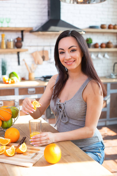 Smiling young woman in a gray T-shirt squeezes a fresh orange on fruit juice. Healthy eating concept. In the interior of the kitchen - Foto, immagini