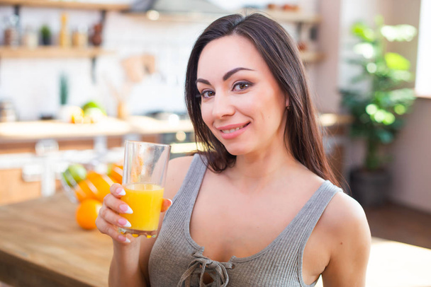 Smiling young woman in gray t-shirt drinks fresh orange juice in kitchen interior - Photo, image