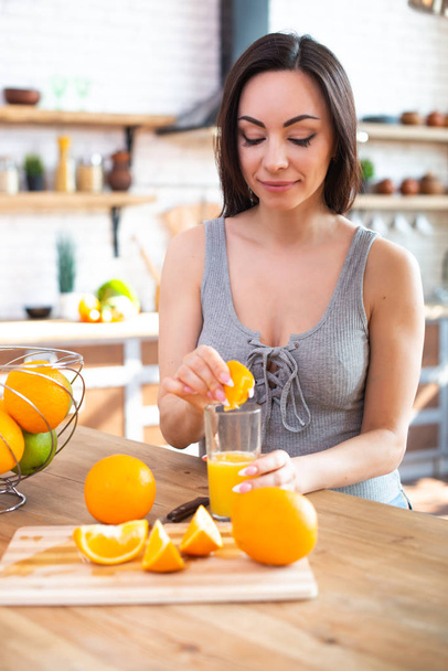 Smiling young woman in a gray T-shirt squeezes a fresh orange on fruit juice. Healthy eating concept. In the interior of the kitchen - Foto, Imagem
