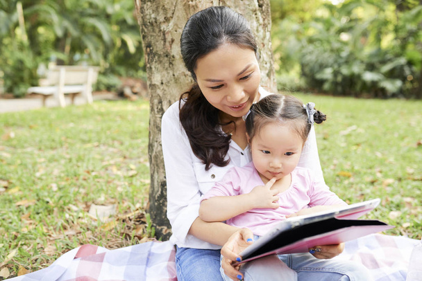 Madre e hija asiáticas sentadas bajo el árbol en el parque de la ciudad y viendo dibujos animados en una tableta digital
 - Foto, imagen