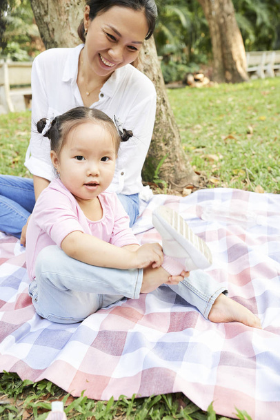 Boldog anya nézett kislánya vesz le cipők, ahogy ül a takaró a városi parkban - Fotó, kép