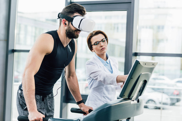 αθληταράς στην προπόνηση με ακουστικά VR στο διάδρομο κοντά στο γιατρό κατά τη διάρκεια της δοκιμής αντοχής στο γυμναστήριο - Φωτογραφία, εικόνα