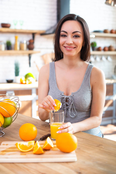 Η χαμογελαστή νεαρή γυναίκα με ένα γκρίζο μπλουζάκι σφίγγει ένα φρέσκο πορτοκάλι στον χυμό φρούτων. Έννοια της υγιεινής διατροφής. Στο εσωτερικό της κουζίνας - Φωτογραφία, εικόνα