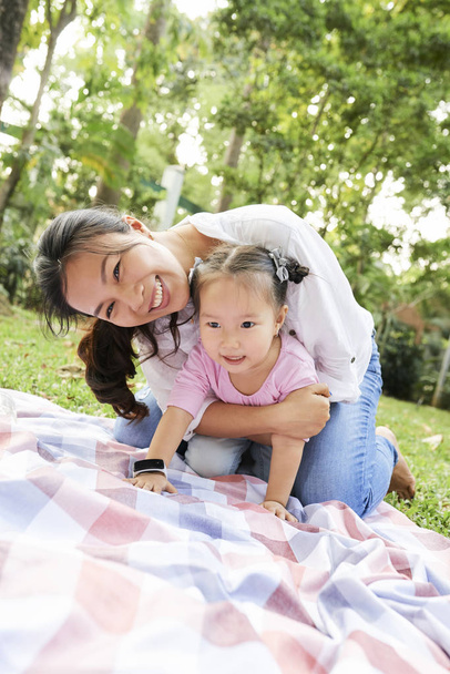 Cheerful Vietnamese mother and daughter playing on blanket in city park - Photo, image