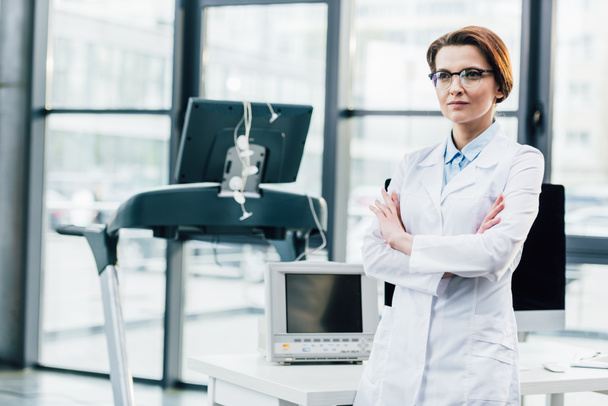 doctor in white coat with crossed arms near treadmill and computer desk at gym - Foto, Bild