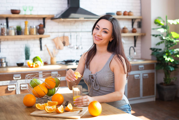 Smiling young woman in a gray T-shirt squeezes a fresh orange on fruit juice. Healthy eating concept. In the interior of the kitchen - Valokuva, kuva