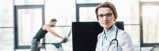 panoramic shot of beautiful doctor in glasses looking at camera near computer in gym - Foto, Bild