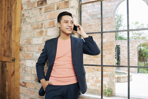 Позитивный красивый предприниматель, стоящий у большого окна в лофт-офисе и разговаривающий по телефону
 - Фото, изображение