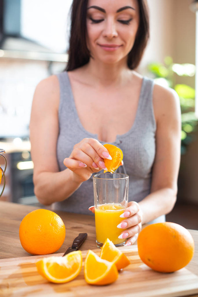 Smiling young woman in a gray T-shirt squeezes a fresh orange on fruit juice. Healthy eating concept. In the interior of the kitchen - Foto, Imagen