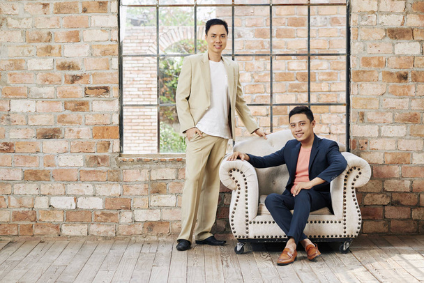 Positive young Asian business partners posing in loft office with brick walls and big windows  - 写真・画像