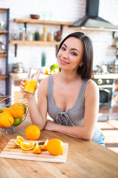 Smiling young woman in gray t-shirt drinks fresh orange juice in kitchen interior - 写真・画像