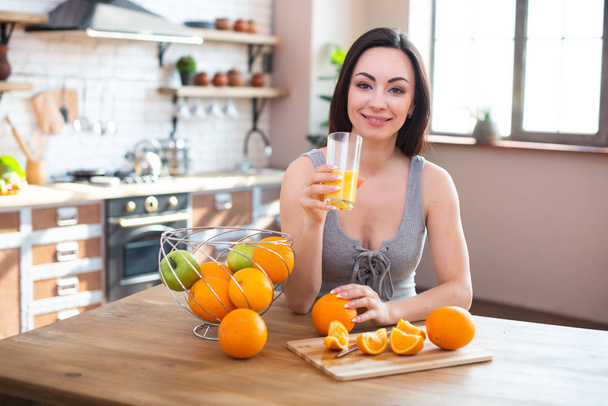Smiling young woman in gray t-shirt drinks fresh orange juice in kitchen interior - Фото, изображение