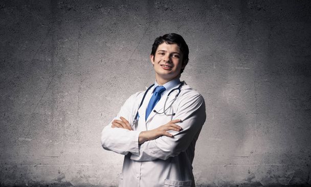 Portrait confident and professional man doctor in white medical suit keeping arms crossed and looking to camera while standing against dark gray wall on background. Medical industry employee concept - 写真・画像