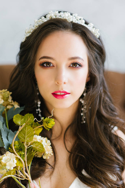 portrait of a beautiful bride with a branch of flowers in her hands. brunette with flowing hair and make-up. morning of the bride. tiara in her hair. cute smiling young girl in a wedding dress - Photo, image