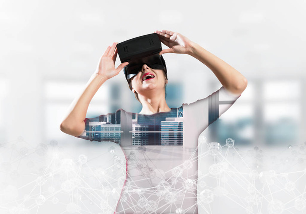 Double exposure of young and beautiful woman in red dress using virtual reality headset while standing indoors of office building with digital network structure. - Фото, изображение