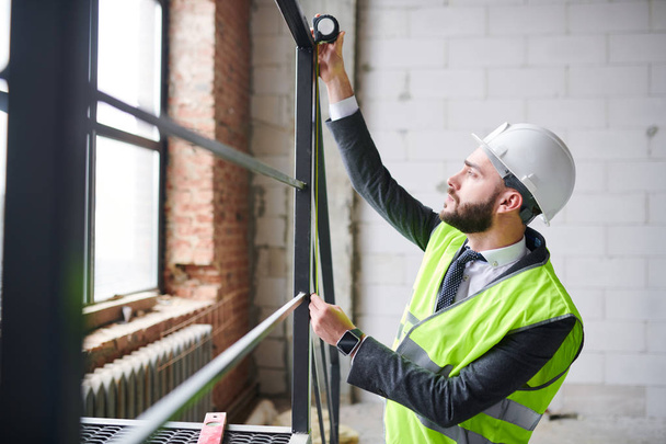 Fiatal kortárs mérnök munkaruhában a fémszerkezet magasságát méri, miközben a befejezetlen épület belsejében dolgozik - Fotó, kép