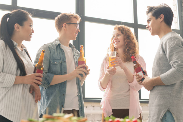 Vier gelukkige jonge casual vrienden die drankjes houden terwijl ze ideeën delen over waar ze moeten gaan in het weekend - Foto, afbeelding