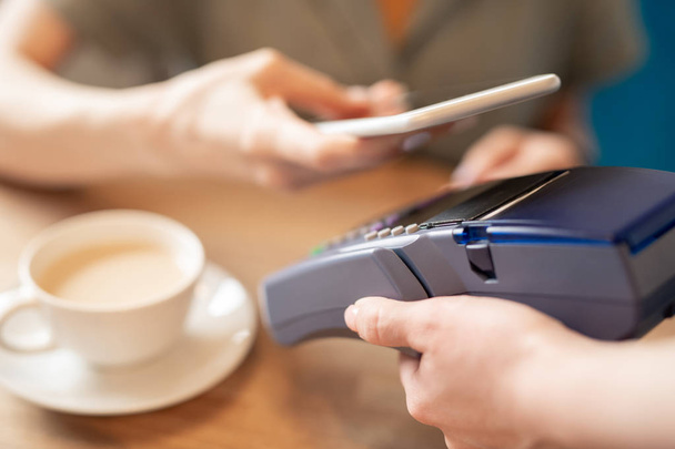 Eigentijds persoon met smartphone over elektronische machine tijdens het gebruik van contactloos betalen in café of restaurant - Foto, afbeelding