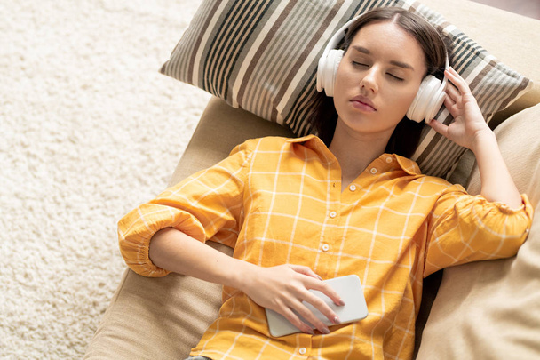 Молода спокійна брюнетка жінка в навушниках лежить на дивані, слухаючи розслаблюючу або її улюблену музику на дозвіллі
 - Фото, зображення
