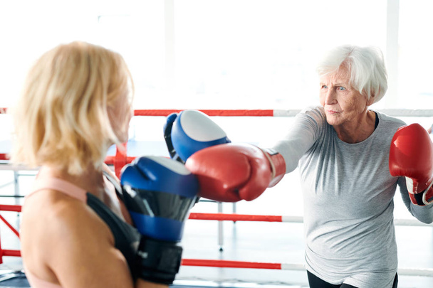 Mujer mayor en ropa deportiva y guantes de boxeo pateando a su joven rival mientras ambos se preparan para la competencia deportiva
 - Foto, imagen