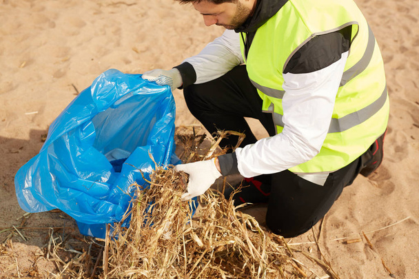 Joven hombre servicio de protección del medio ambiente voluntario limpieza playa y poner basura en azul saco
 - Foto, Imagen