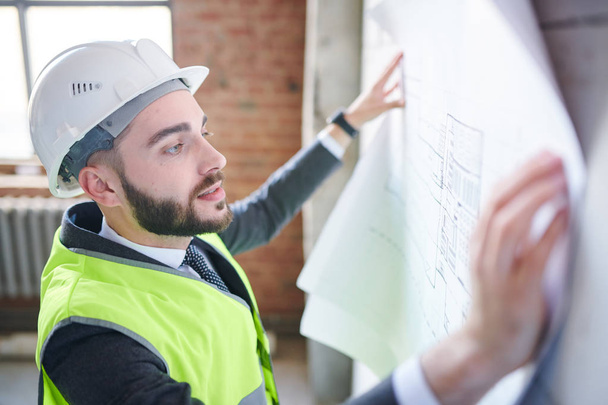 Young engineer in hardhat and uniform looking at sketch on blueprint while standing in front of wall of unfinished building - Foto, Imagem