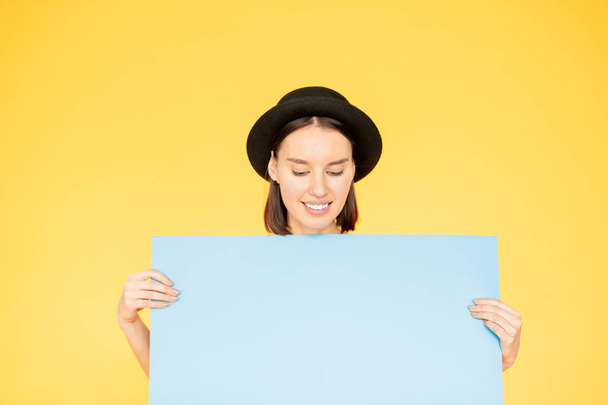 Young smiling female in black hat looking at blue sheet in her hands while making advert on yellow background - Foto, afbeelding