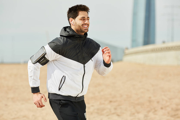 Młody, wesoły sportowiec w odzieży sportowej biegający po plaży w słoneczny poranek słuchając ulubionej muzyki - Zdjęcie, obraz