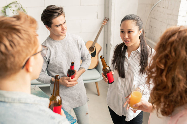 Pretty mixed-race brunette with bottle of beer looking at one of guys while explaining her idea to friends - Photo, Image