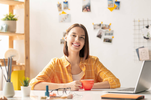 Happy brunette girl in casualwear looking at you with toothy smile while sitting by desk in office, having drink and networking - Foto, afbeelding