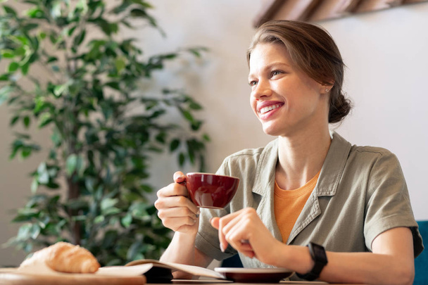 Jovem mulher morena alegre com sorriso de dente desfrutando de pausa no café enquanto se senta à mesa e toma uma xícara de café
 - Foto, Imagem