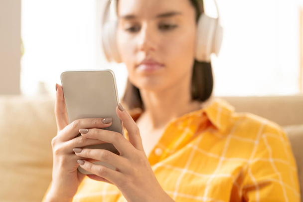 Mãos de jovem mulher contemporânea em fones de ouvido segurando smartphone enquanto percorre a lista de reprodução ou mensagens de texto
 - Foto, Imagem