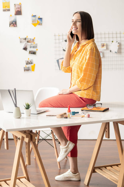 Szczęśliwy młody ładny projektant w casualwear rozmawia na telefon komórkowy podczas posiedzenia na biurku przed laptopem - Zdjęcie, obraz
