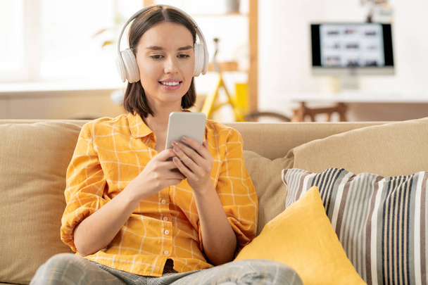 Mujer morena joven feliz en ropa de casualwear sentado en el sofá mientras escucha música en los auriculares y desplazamiento en el teléfono inteligente
 - Foto, imagen