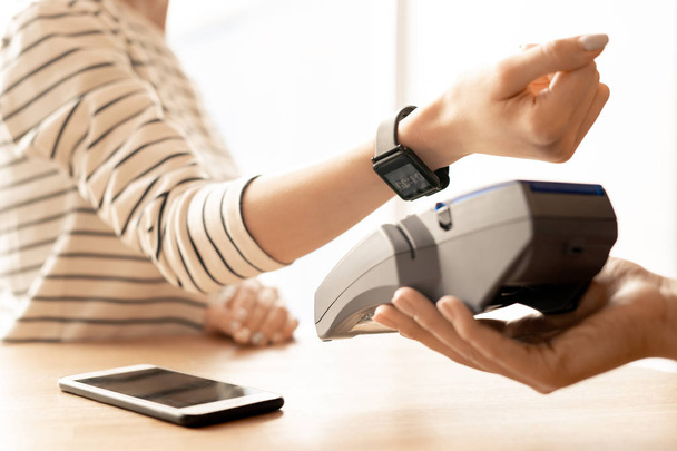 Рука молодой современной женщины с умными часами на запястье, держащей его над электронной машиной при оплате товаров или услуг
 - Фото, изображение