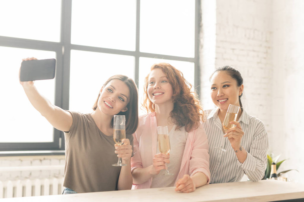 Τρία ευτυχισμένα κορίτσια σε ανεπίσημο ντύσιμο φτιάχνοντας selfie ενώ έχουν σαμπάνια κατά τη διάρκεια του εορτασμού της ζωής εκδήλωση - Φωτογραφία, εικόνα