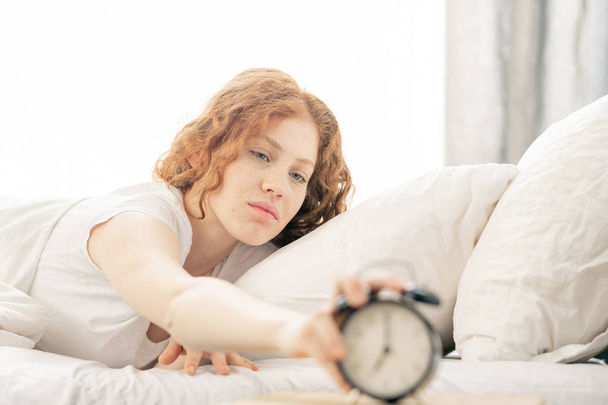 Displeased young woman lying on bed while stretching hand to turn off the alarm clock in the morning - Photo, image