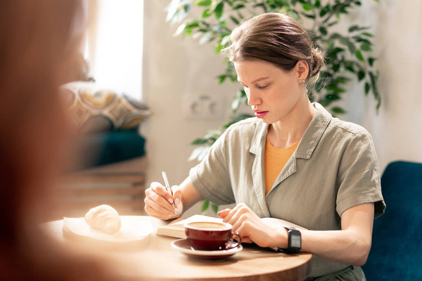 Mladá současná obchodnice v casualwear sedí u stolu v kavárně, dává si kávu s pečivem a dělá si poznámky do zápisníku - Fotografie, Obrázek
