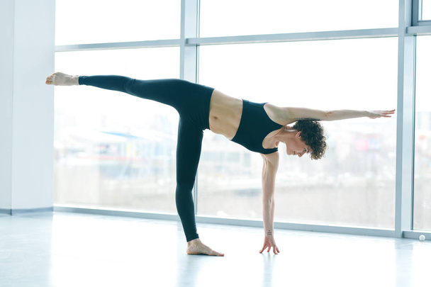 Young active sportswoman standing on left leg and arm while stretching the right ones and bending over floor during workout - Фото, зображення