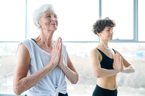 Aktywna Starsza kobieta w odzieży sportowej trzymając ręce połączone przez klatkę piersiową, ćwicząc relaksujące ćwiczenia jogi w siłowni - Zdjęcie, obraz
