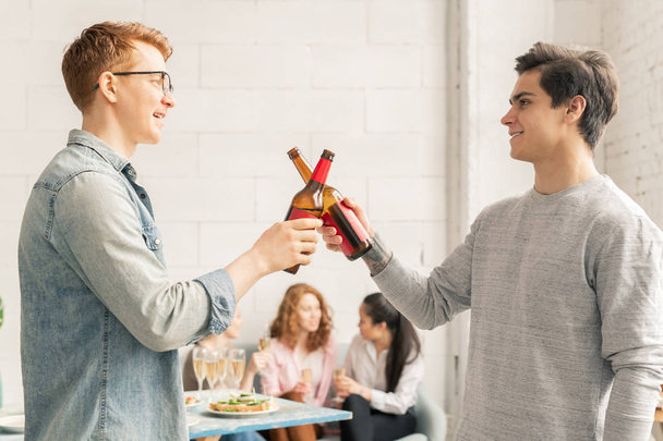 Двоє молодих випадкових чоловіків смердить пляшками пива на домашній вечірці, роблячи тости на фоні дівчат з шампанським
 - Фото, зображення