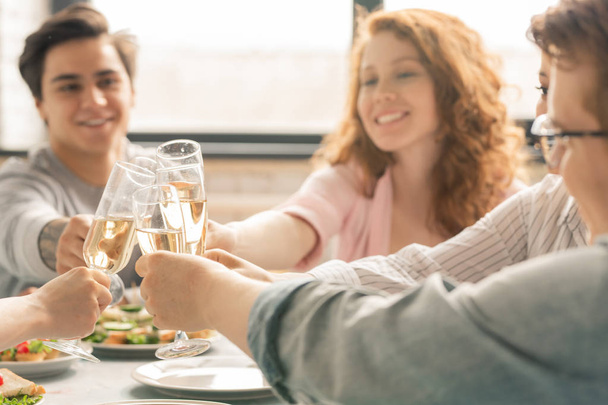 Group of friends clinking with flutes of champagne over homemade snacks on served table while celebrating life event - 写真・画像